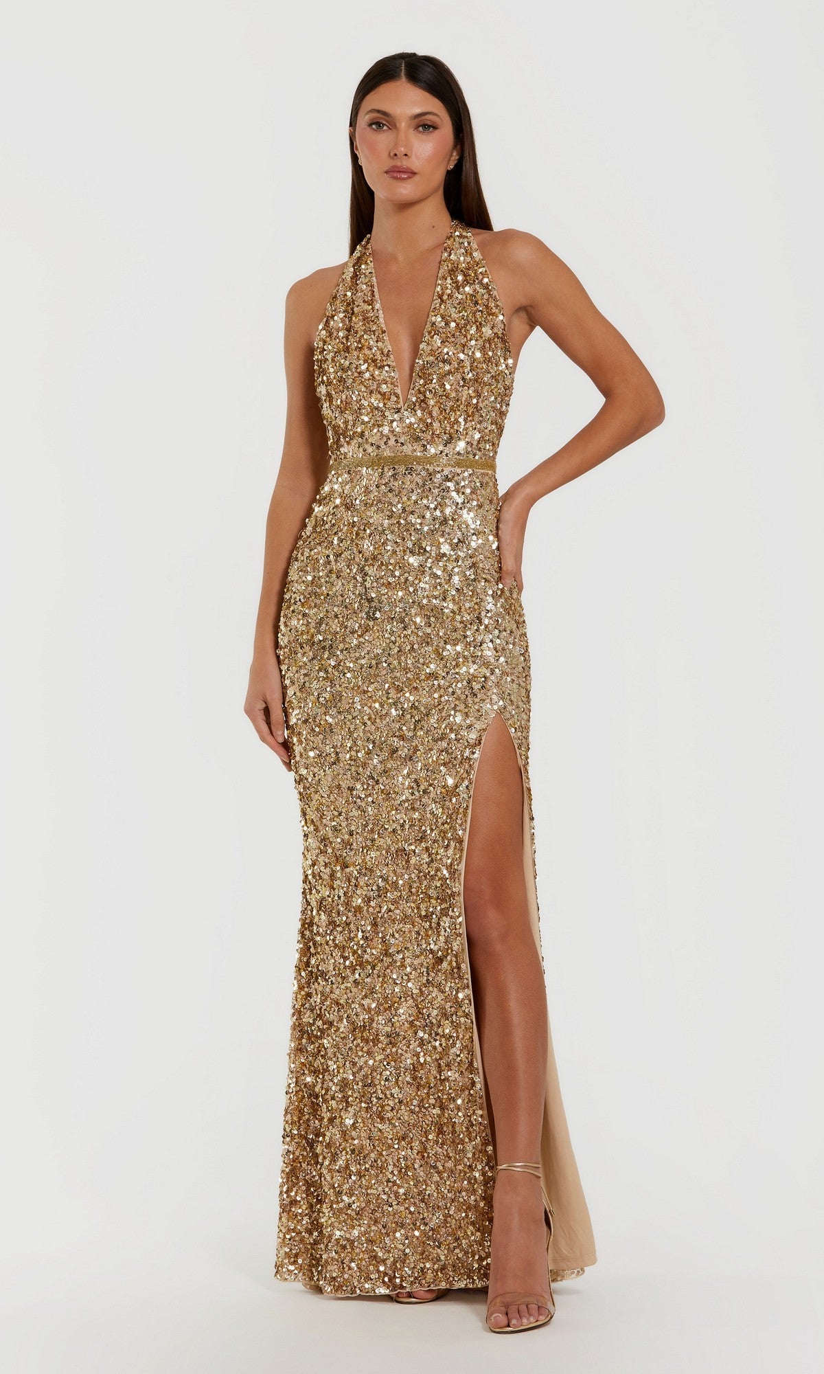 A woman stands against a plain white background wearing the Mac Duggal 93745 long formal dress in gold, featuring allover sequins, a halter neckline with a deep V front, fitted waist, and thigh-high slit, paired with nude heels.